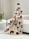 Woman in a cream floral printed abaya, holding a taupe clutch, seated indoors
