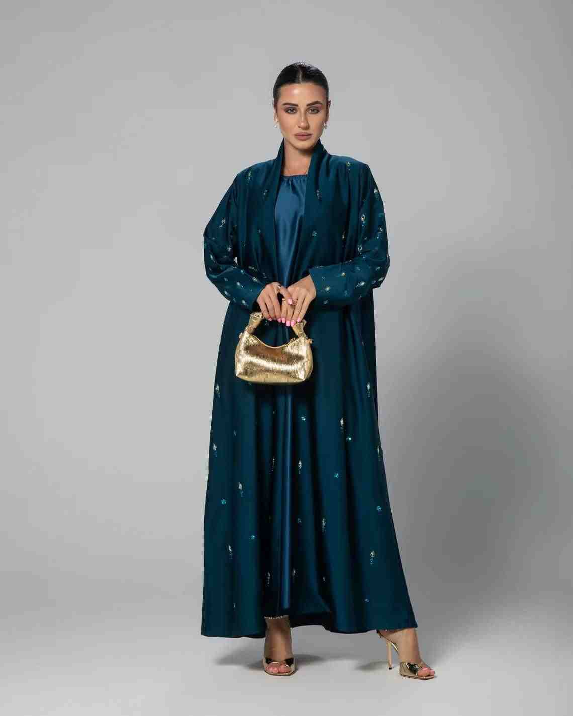Woman in teal designer abaya with gold accents holding a gold handbag, standing in studio