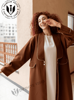 Woman in a brown designer abaya with white trim, standing on a balcony in daylight