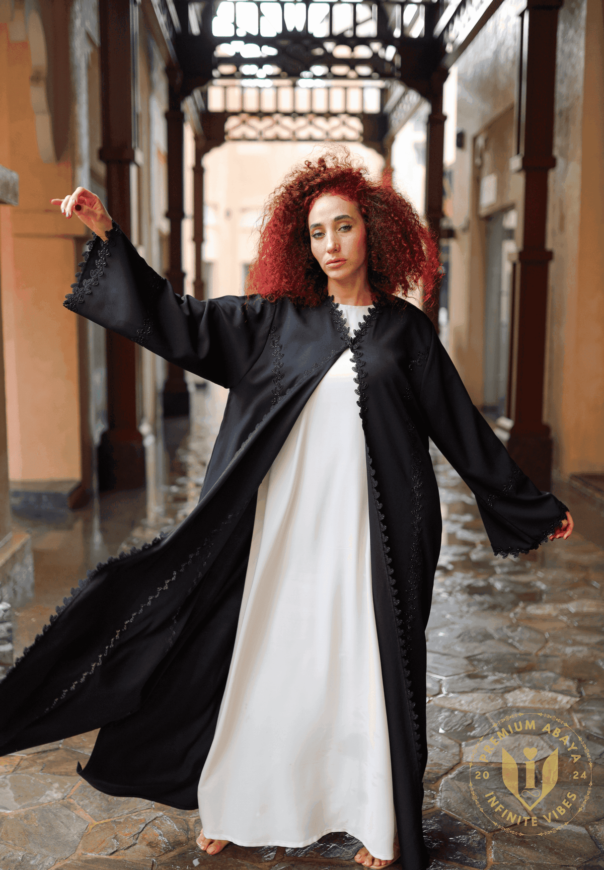 Woman wearing a black embroidered designer abaya over a white dress, standing in a traditional UAE alleyway