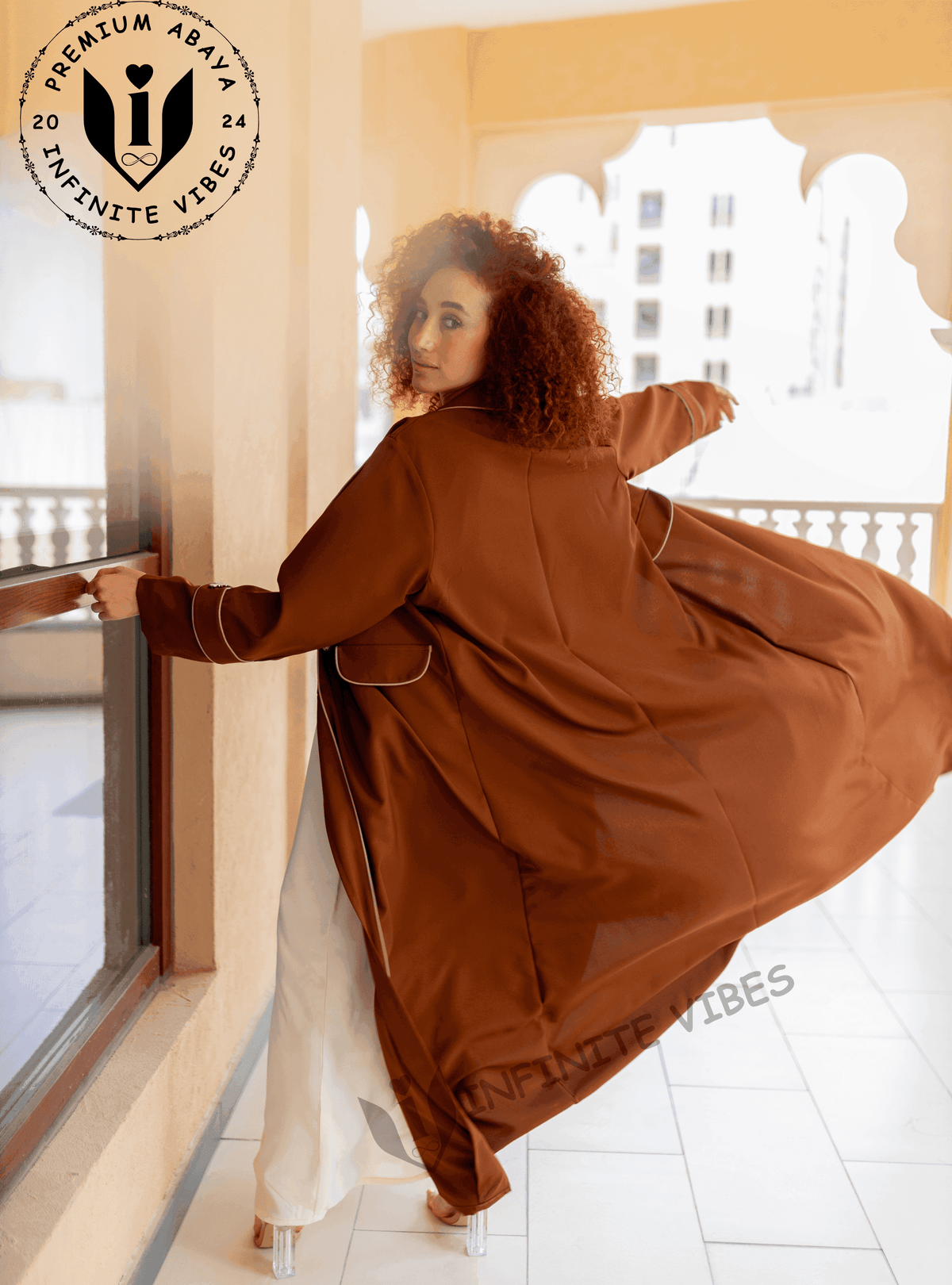 Woman in flowing brown designer abaya with white trim, standing on balcony, modest fashion.