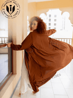 Woman in flowing brown designer abaya with white trim, standing on balcony, modest fashion.