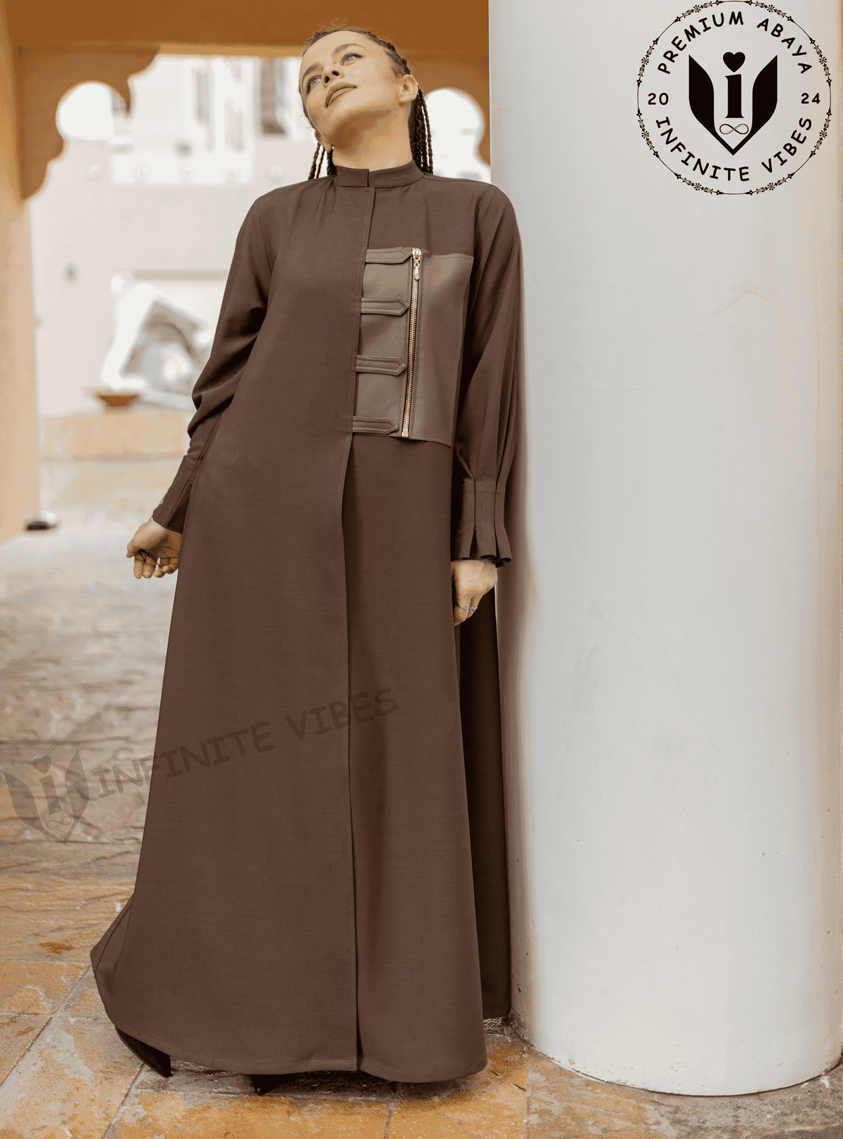 Woman in brown designer abaya with zip detail, standing outdoors by a column.