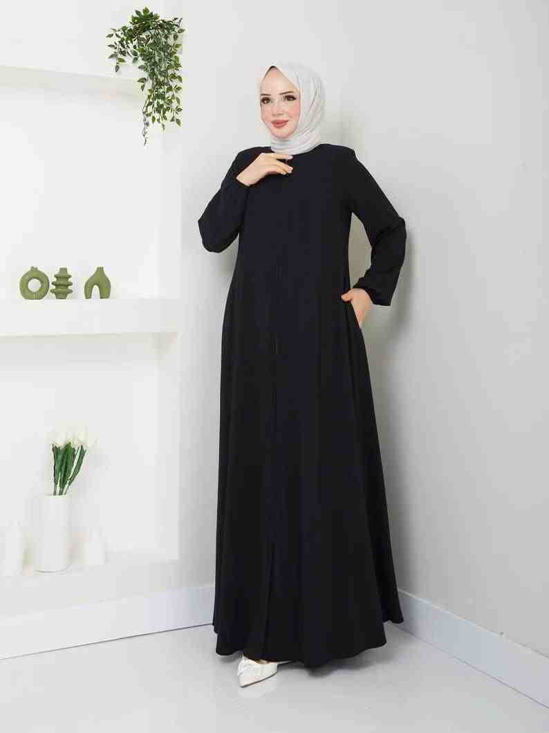 Woman in a simple black abaya and white hijab standing in a modern minimal room.