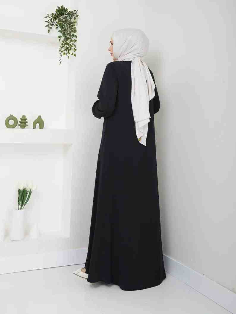 Woman wearing a black abaya and light hijab, standing in minimal modern room