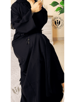 Elegant black abaya with subtle leaf embroidery, worn indoors on a light modern background.