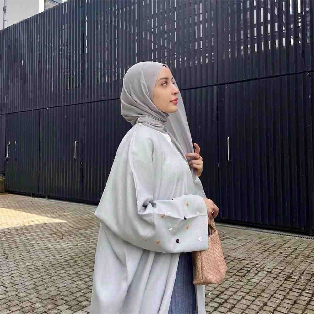 Elegant woman in light grey embroidered abaya and hijab outdoors, holding a woven handbag