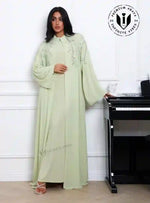 Woman wearing premium pistachio green chiffon abaya with crystal vine embroidery, standing indoors