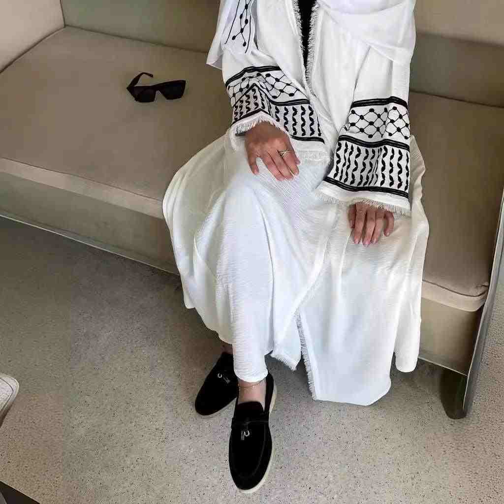 Woman wearing white abaya with black keffiyeh embroidery and tassels, seated indoors.