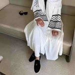 Woman wearing white abaya with black keffiyeh embroidery and tassels, seated indoors.