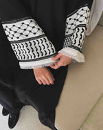 Black abaya with white keffiyeh-patterned embroidered sleeves and tassel trim.