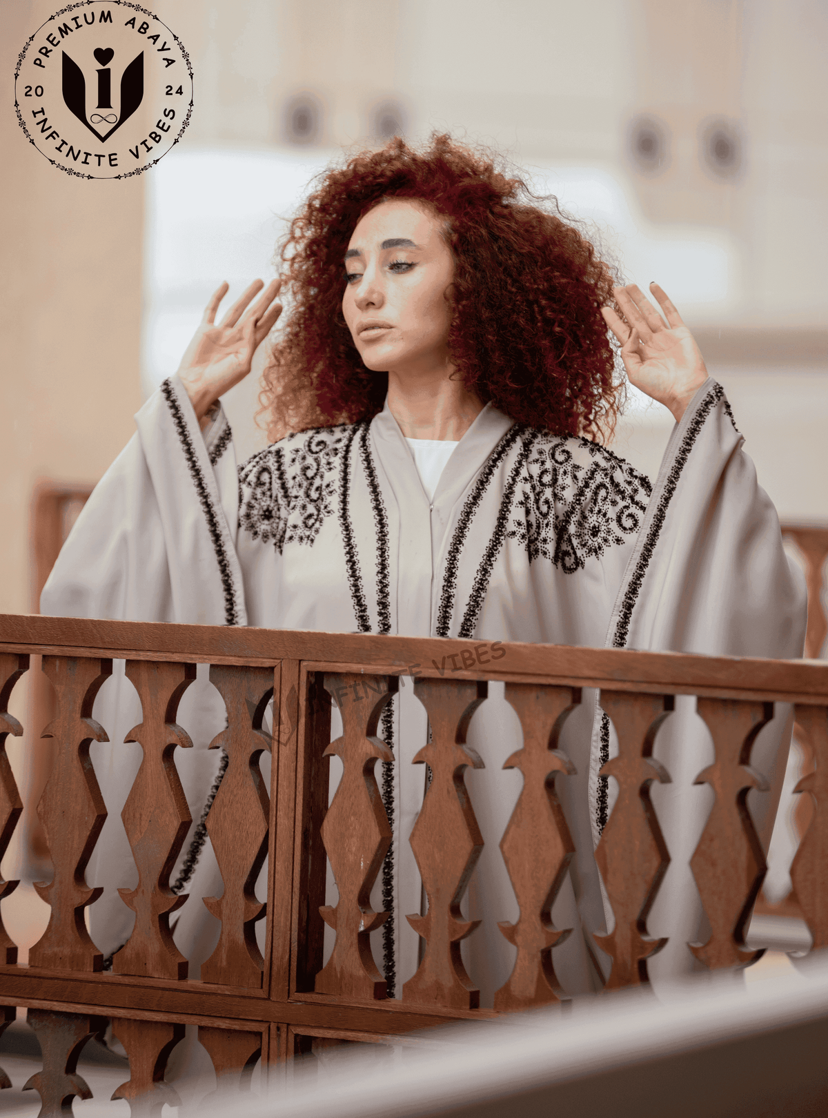 Woman in light grey butterfly abaya with black embroidery, standing by wooden lattice