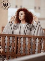 Woman in light grey butterfly abaya with black embroidery, standing by wooden lattice
