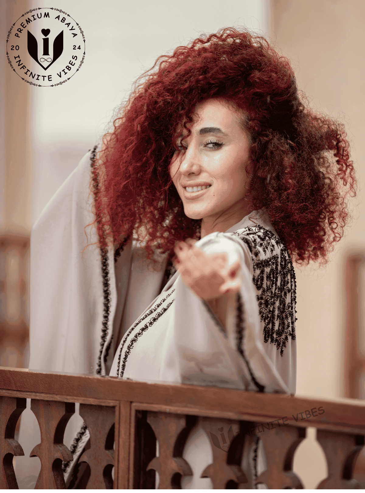 Woman with curly red hair in an embroidered butterfly abaya, smiling on a balcony.