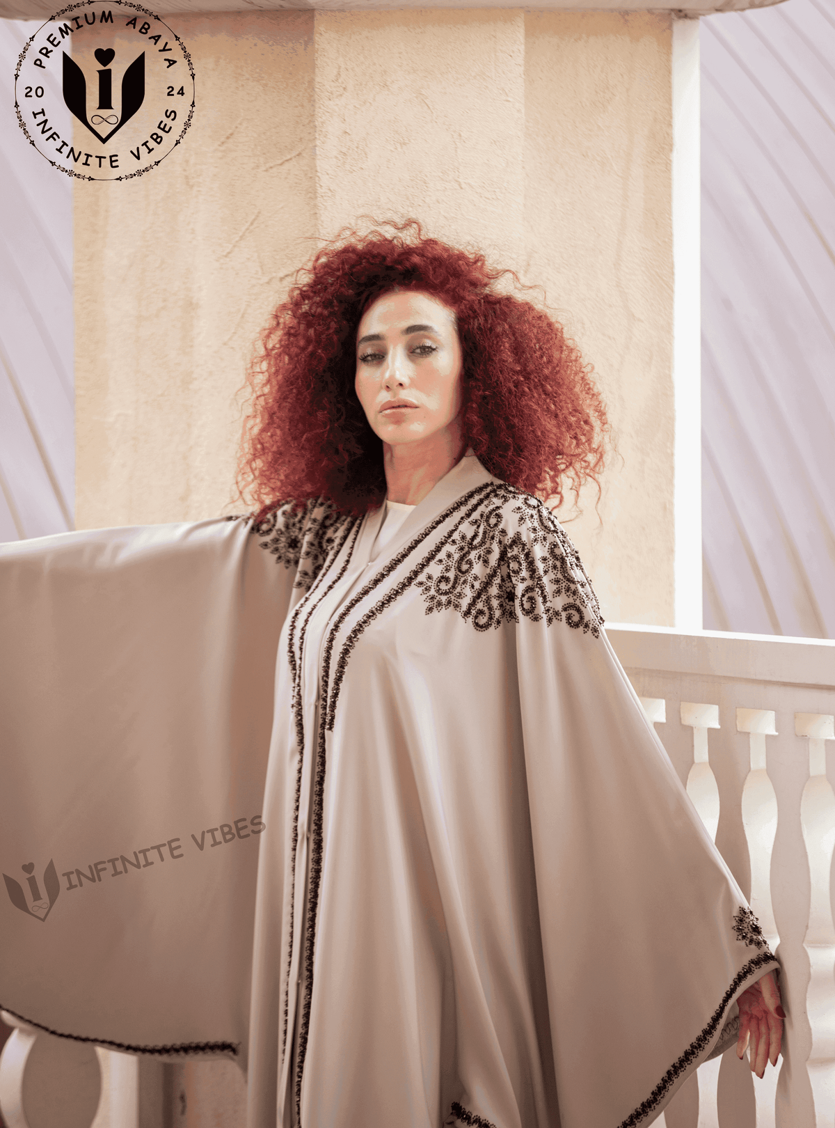 Woman wearing a butterfly abaya with black embroidery, standing by a cream balcony.