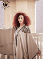 Woman wearing a butterfly abaya with black embroidery, standing by a cream balcony.