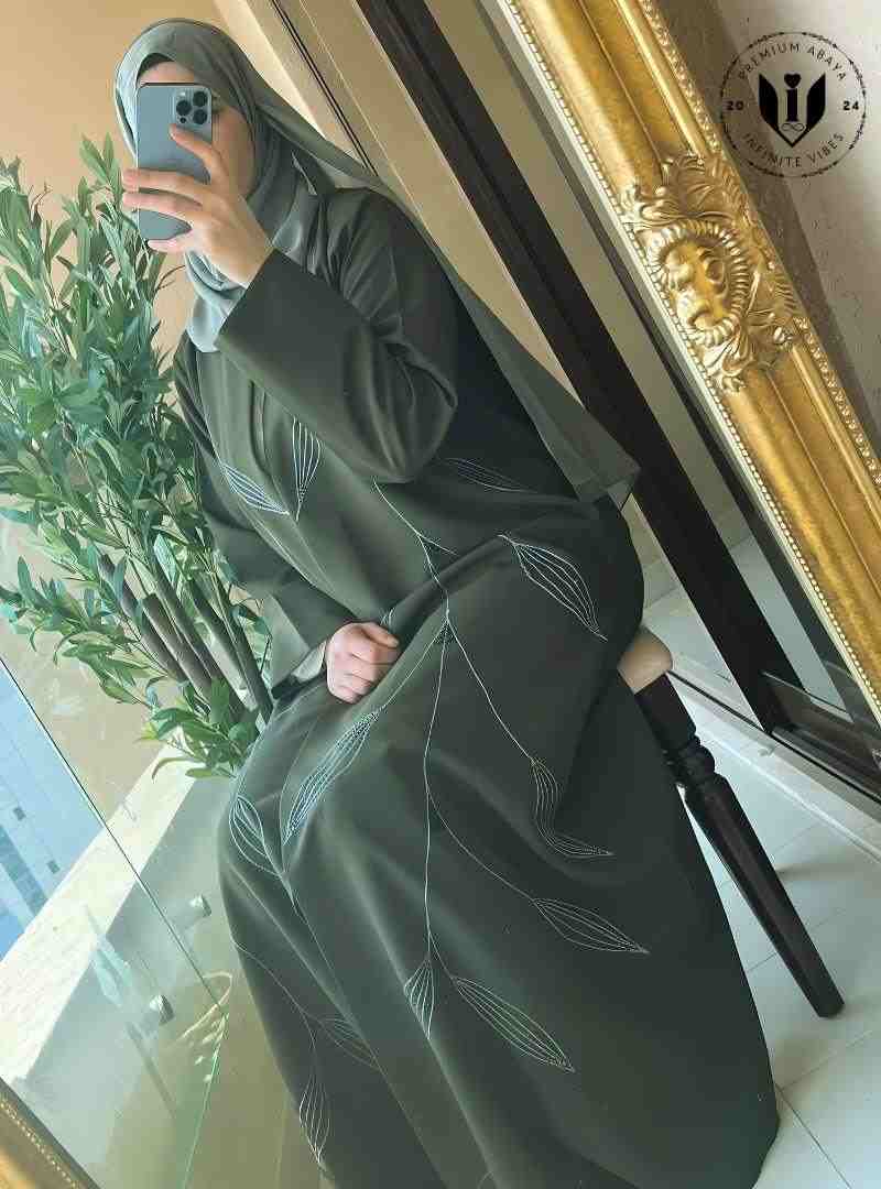 Woman in modern olive green abaya with leaf embroidery, sitting by a mirror indoors