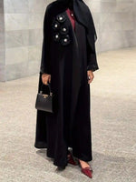 Elegant black abaya with floral embellishments, paired with a black handbag and burgundy flats.
