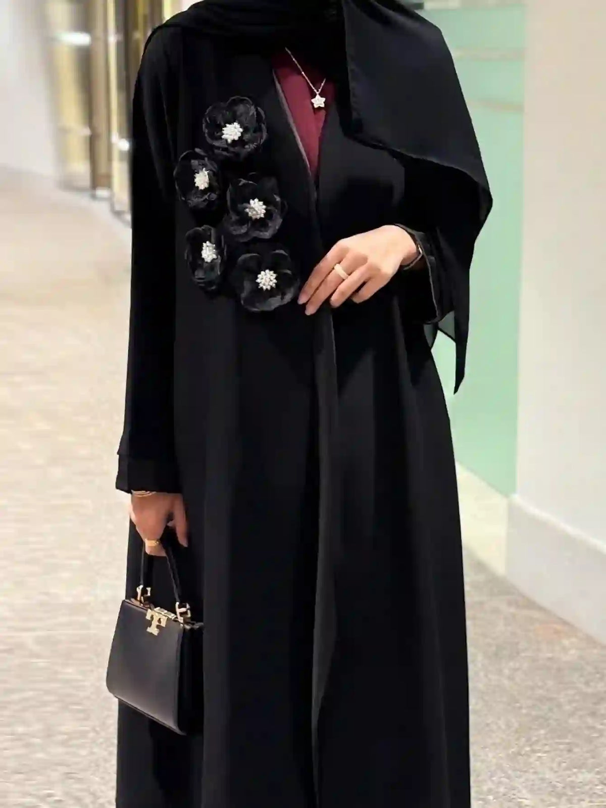 Modern black abaya with floral embellishments, paired with a small black handbag