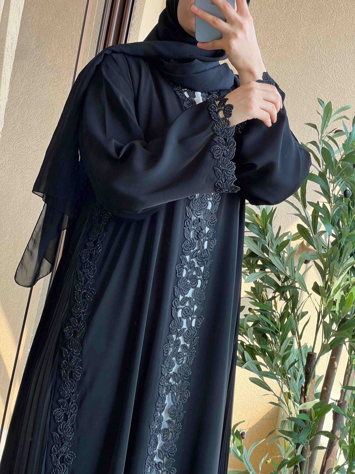 Elegant black abaya with intricate lace embroidery, worn by woman indoors near plant