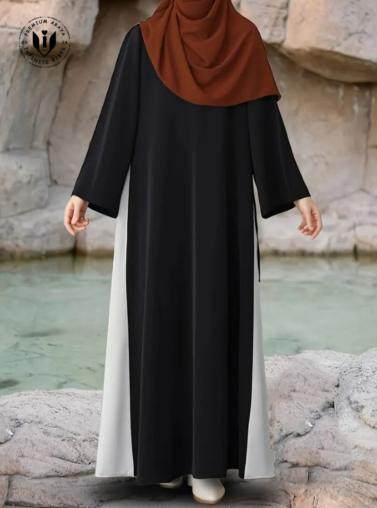 Person wearing a black and white abaya with a brown hijab against a stone wall and water background