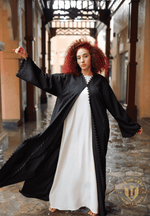 Woman wearing a black embroidered designer abaya over a white dress, standing in a traditional UAE alleyway