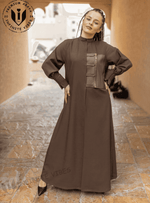 Woman wearing a brown abaya with front zip detail, standing in a modern outdoor corridor.