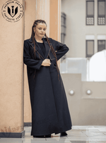 Woman wearing a black designer abaya with gold trim, standing outdoors in a modern setting