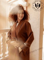 Woman wearing a brown designer abaya with white trim, standing on a sunlit balcony