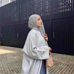 Elegant woman in light grey embroidered abaya and hijab outdoors, holding a woven handbag