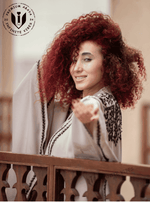 Woman with curly red hair in an embroidered butterfly abaya, smiling on a balcony.
