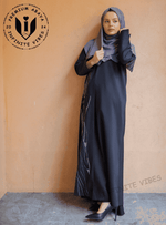 Woman in black handwork abaya with silver embroidery, wearing grey hijab, standing indoors