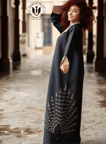 Woman wearing a black embroidered abaya with gold detailing in an outdoor corridor setting.