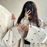 Women wearing cream abayas with colorful heart embroidery, making a heart hand gesture.