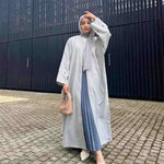 Woman wearing light grey embroidered open abaya, hijab, and pleated skirt, holding a handbag.