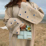 Beige abaya with blue heart embroidery, wide sleeves, and light blue handbag on beach