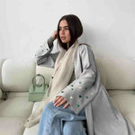 Woman in light grey open abaya with green heart embroidery, sitting on a white sofa